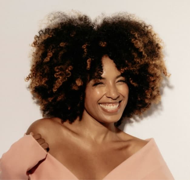 A woman with a full afro in a blush off-shoulder top, smiling